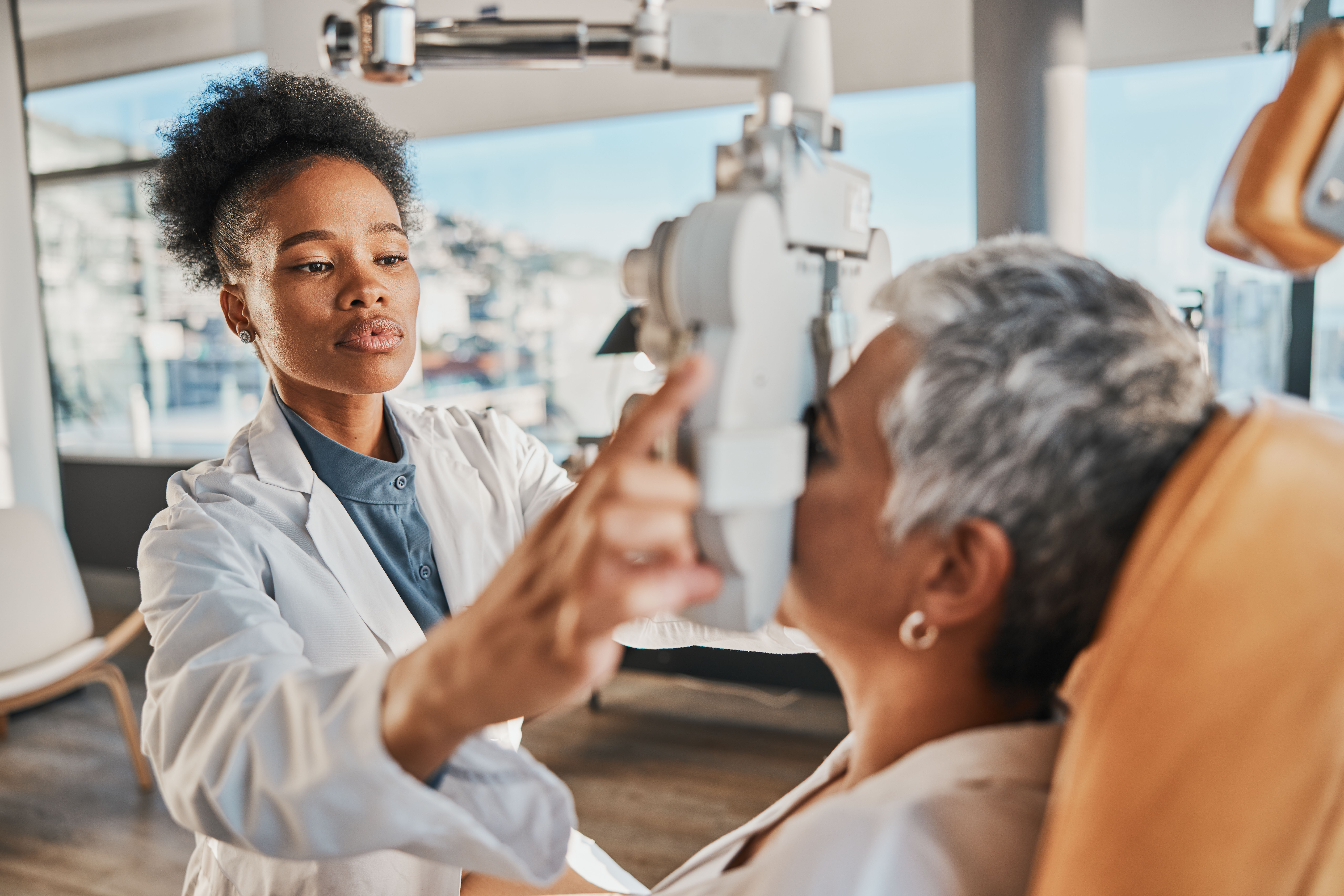 An optometrist, wearing a white coat and focused expression, operates an eye examination device while assisting an older patient with short gray hair seated in a medical chair. The scene is set in a modern, sunlit clinic.